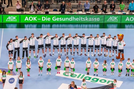 Eine Männerhandballmannschaft in weißen Jacken steht in einer Reihe, die Arme umeinander gelegt, neben einem Maskottchen. Kinder in grün-weißen Uniformen stehen davor. Im Hintergrund sind Zuschauer und eine große grüne Fahne zu sehen.