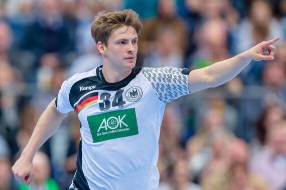 Ein männlicher Handballspieler in einem weiß-schwarzen Trikot mit der Nummer 34 zeigt während eines Spiels nach vorne, mit einer unscharfen Menschenmenge im Hintergrund.