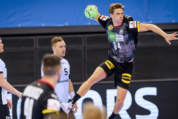 Ein Handballspieler in einem schwarz-grauen Trikot springt in der Luft, um einen grünen Ball zu werfen, während drei Verteidiger in weißen Trikots ihn während eines Spiels beobachten.
