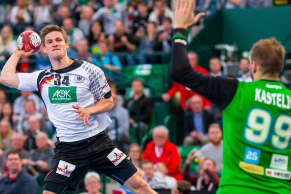 Ein männlicher Handballspieler in einem weiß-schwarzen Trikot bereitet sich darauf vor, den Ball zu werfen, während ein Torwart in einem grünen Trikot mit der Kastelic 99 die Hände zum Abwehren vor einer jubelnden Menge hebt.