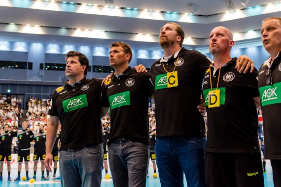 Fünf Männer in passenden schwarzen Hemden mit grünem Logo stehen Arm in Arm mit dem Gesicht nach vorne in einer Halle. Im Hintergrund steht eine Mannschaft, und das Publikum füllt die Tribünen. Der Schauplatz scheint eine Sportveranstaltung zu sein.