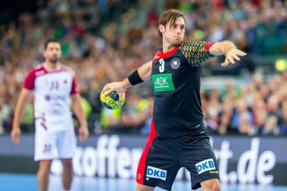 Ein männlicher Handballspieler in einem schwarz-roten Trikot, der einen grün-gelben Ball in der Hand hält, bereitet sich auf den Wurf während eines Spiels vor. Ein anderer Spieler in einer weißen Uniform steht im Hintergrund auf einem überfüllten Hallenplatz.