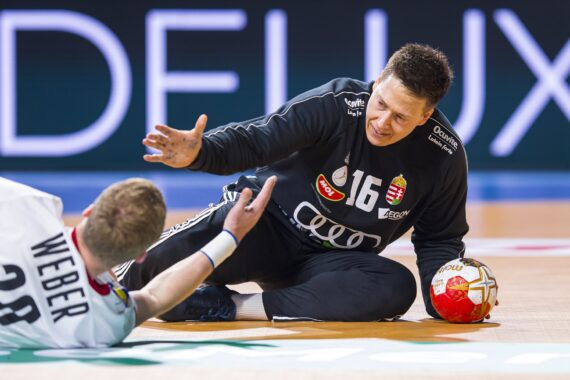 Ein Handballtorwart in einem schwarzen Trikot sitzt auf dem Spielfeld, hält den Ball und streckt die Hand nach einem Spieler in einem weißen Trikot mit der Aufschrift Weber 28 aus, der auf dem Boden liegt.