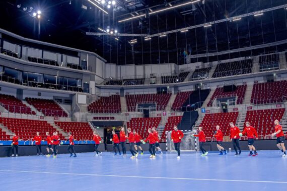 Eine Gruppe von Sportlern in roten Hemden trainiert auf einem großen Hallenplatz mit blauem Bodenbelag in einer leeren Arena mit roten und grauen Sitzen. Helle Lichter erhellen die geräumige, moderne Halle.