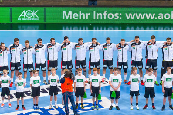 Eine Herrenhandballmannschaft in weißen und schwarzen Trainingsanzügen steht in einer Reihe und legt die Arme umeinander. Vor ihnen stehen Kinder in weißen Hemden nebeneinander auf einem blauen Spielfeld. Auf der linken Seite steht ein Maskottchen.