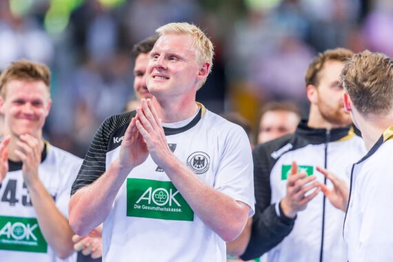 Ein männlicher Handballspieler in einem weiß-schwarzen Trikot klatscht und lächelt nach einem Spiel, umgeben von Mannschaftskameraden. Die Mannschaft trägt weiße Trikots mit grünen Sponsorenlogos. Der Hintergrund ist unscharf und es sind mehrere Personen zu sehen.