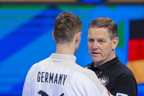 Ein Trainer und ein Spieler, der ein Deutschland-Trikot mit Puma-Logo trägt, unterhalten sich während einer Sportveranstaltung, wobei die deutsche Flagge im Hintergrund verschwommen zu sehen ist.