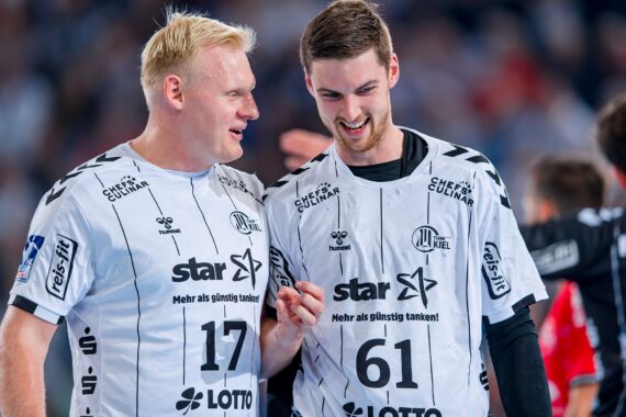 Zwei männliche Handballspieler in weißen Trikots mit den Nummern 17 und 61 stehen eng beieinander, lächeln und unterhalten sich während eines Spiels. Der Hintergrund ist unscharf und zeigt andere Spieler und eine jubelnde Menge.