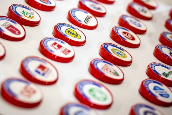 Auf einer weißen Fläche sind Reihen von runden Ansteckern oder Pins mit verschiedenen farbigen Logos und rot umrandeten Texten angebracht. Die Anstecknadeln stehen für verschiedene Organisationen oder Institutionen.