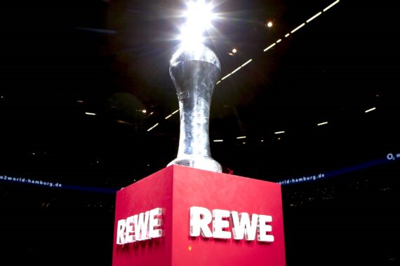 Auf einem roten Sockel steht eine silbern glänzende Trophäe mit dem Schriftzug REWE in weißen Buchstaben, der sich dramatisch von einem dunklen Hintergrund abhebt.