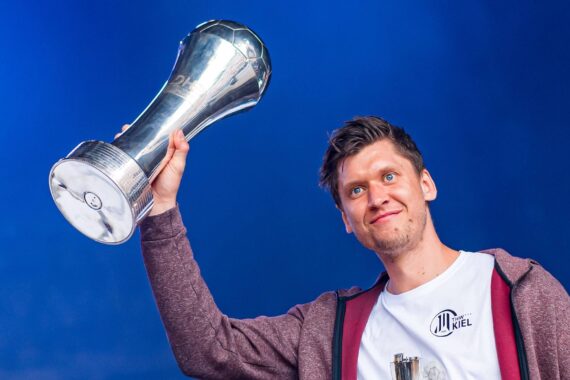 Ein Mann mit kastanienbraunem Kapuzenpulli und weißem T-Shirt hält mit einer Hand lächelnd einen großen silbernen Pokal vor einem blauen Hintergrund hoch.