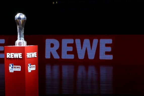 Ein silberner Sportpokal steht auf einem roten Sockel mit REWE-Branding vor einem großen beleuchteten REWE-Schild auf dunklem Hintergrund.