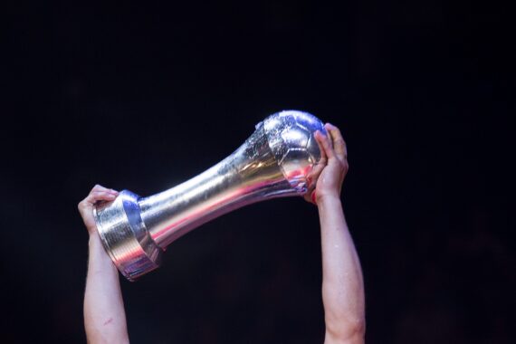 Eine Person hält mit beiden Händen eine große silberne Trophäe vor einem dunklen Hintergrund in die Höhe und feiert einen Sieg.