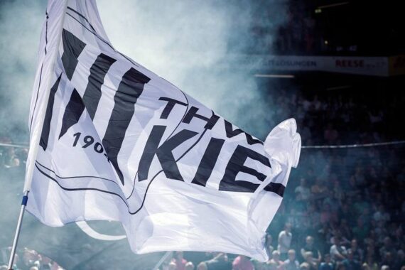Eine große weiße Flagge mit THW KIEL und 1904 in schwarzer Schrift weht in einer verrauchten, überfüllten Arena während einer Sportveranstaltung.