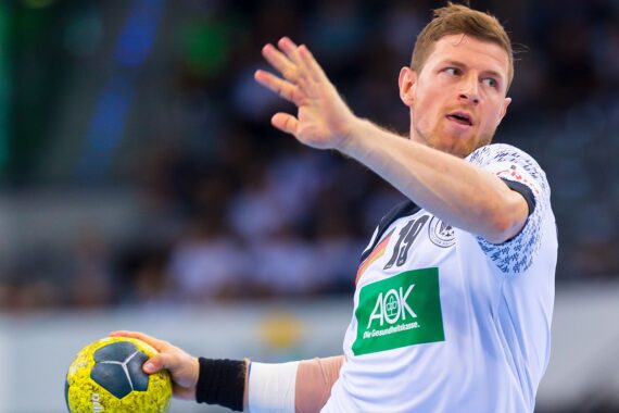 Ein männlicher Handballspieler in einem weißen Trikot bereitet sich darauf vor, einen gelben und blauen Ball während eines Spiels zu werfen, mit konzentriertem Gesichtsausdruck und verschwommenen Zuschauern im Hintergrund.