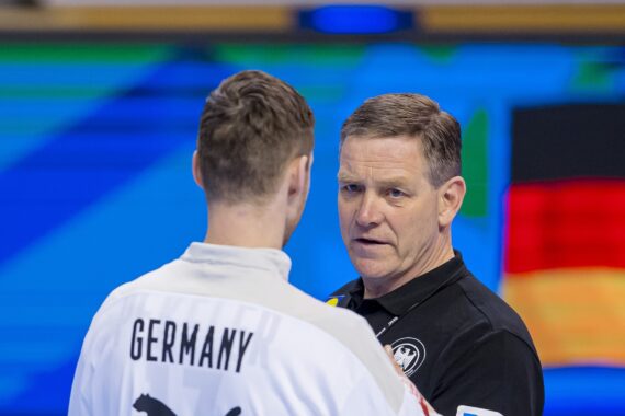 Ein Mann, der eine deutsche Sportjacke trägt, spricht mit einem Trainer oder Beamten in einem schwarzen Hemd. Im Hintergrund sind die deutsche Flagge und ein blauer Hintergrund zu sehen.