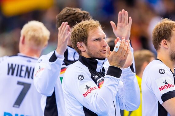 Mehrere männliche Athleten in weißen Sportuniformen klatschen und schauen nach vorne, als ob sie etwas feiern oder anerkennen wollten. Ein Mann mit bandagierten Fingern steht auffällig in der Mitte. Der Fokus liegt auf ihren Oberkörpern.