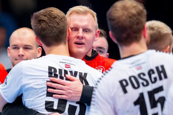 Eine Gruppe männlicher Handballspieler umarmt sich nach einem Spiel, wobei ein Spieler in Rot einen Spieler in einem weißen Trikot mit der Aufschrift "BINK" umarmt, während auf dem Trikot eines anderen Spielers "POSCH" steht. Die Atmosphäre scheint emotional und unterstützend zu sein.