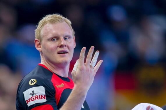 Ein männlicher Sportler in einem schwarz-roten Sporttrikot hält seine linke Hand mit abgeklebten Fingern hoch und blickt in die Kamera. Der Hintergrund ist unscharf, was auf eine Indoor-Sportveranstaltung hindeutet.