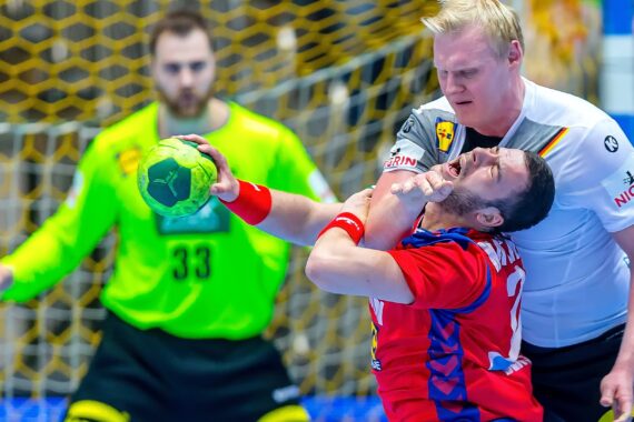 Ein Handballspieler in einem weißen Trikot greift einem Gegner in einem roten Trikot während eines Spiels kräftig ins Gesicht, während ein Torwart in einem neongrünen Trikot vom Torraum aus im Hintergrund zusieht.