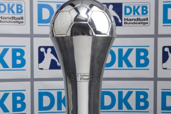Eine silberne Trophäe in Form eines Fußballs auf einem hohen Sockel, der vor einem Hintergrund mit mehreren blauen DKB-Logos und Silhouetten von Handballspielern steht.