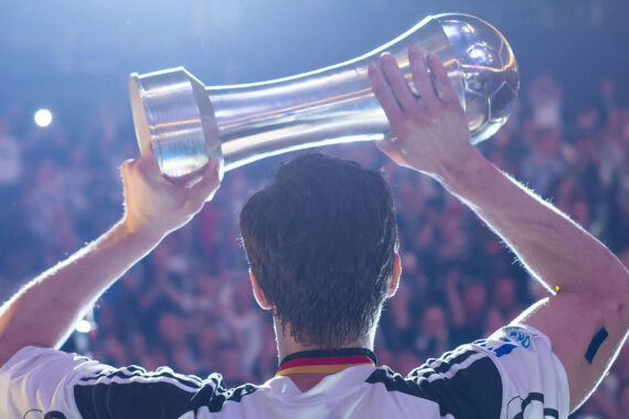Ein Fußballspieler, der von hinten zu sehen ist, hebt eine glänzende Trophäe über seinen Kopf, während im Hintergrund eine jubelnde Menge zusieht.