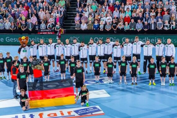 Eine Herren-Sportmannschaft steht in einer Reihe auf einem blauen Spielfeld, trägt weiße Jacken und Kinder in schwarzer Kleidung, die eine große deutsche Flagge vor sich halten. Eine Menschenmenge schaut von den hinteren Tribünen zu.