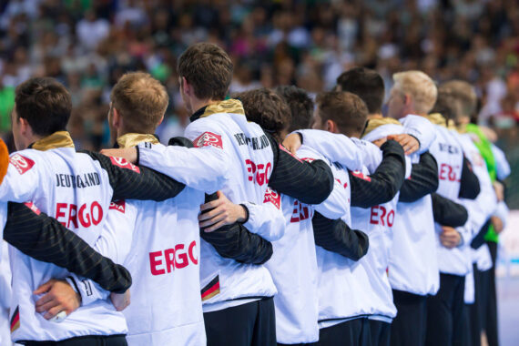 Eine Reihe von Sportlern, die weiße Jacken mit den Aufschriften Deutschland und ERGO tragen, stehen nebeneinander, die Arme um die Schultern gelegt, vor einer Menschenmenge in einer Halle.