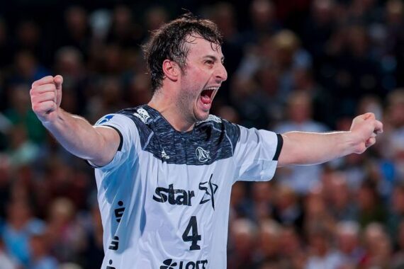 Ein männlicher Handballspieler in einem weiß-schwarzen Trikot mit der Nummer 4 jubelt mit ausgestreckten Armen und offenem Mund, er wirkt aufgeregt und triumphierend während eines Spiels. Die Menge ist im Hintergrund verschwommen.