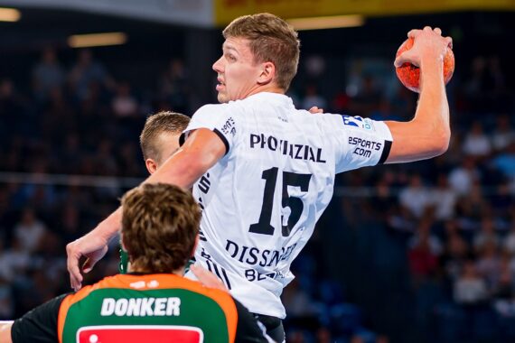 Ein Handballspieler im weißen Trikot bereitet sich auf den Wurf vor und springt während eines intensiven Spiels über zwei gegnerische Spieler in grünen und orangenen Trikots.