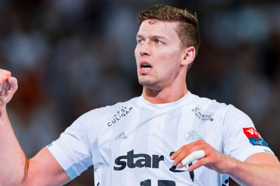 Ein männlicher Handballspieler in einem weißen Trikot feiert mit einer geballten Faust und sieht entschlossen aus. Der Hintergrund ist unscharf, um die Aufmerksamkeit auf den Ausdruck und die Geste des Sportlers zu lenken.