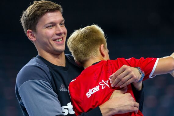 Ein lächelnder Mann in Sportkleidung umarmt einen kleinen Jungen in einem roten Trikot, der einen bunten Handball in der Hand hält, auf einem Hallenplatz. Der Hintergrund ist unscharf.