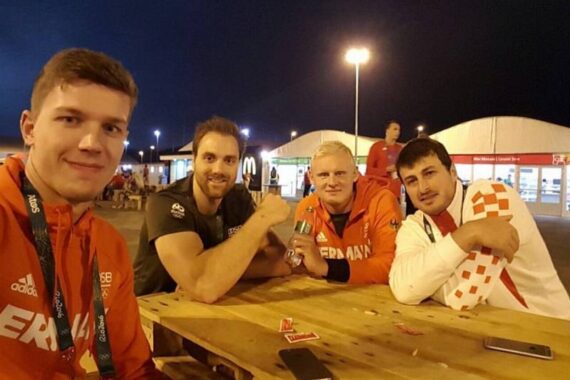 Vier Männer sitzen nachts im Freien um einen Holztisch und lächeln in die Kamera. Sie tragen Sportjacken mit der Aufschrift GERMANY und einem rot-weißen Karomuster, wahrscheinlich Sportler bei einer Veranstaltung. Im Hintergrund sind helle Lichter und Zelte zu sehen.