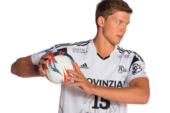 Ein männlicher Handballspieler in einem weißen Trikot mit der Nummer 15 hält einen Handball und bereitet sich darauf vor, ihn zu werfen. Er blickt konzentriert nach links, vor einem schlichten weißen Hintergrund.