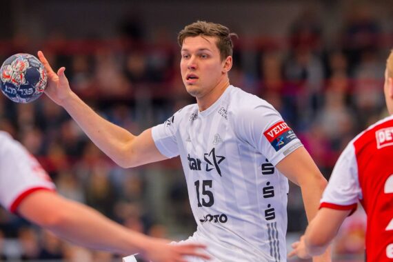 Ein männlicher Handballspieler in einem weißen Trikot mit der Nummer 15 hält einen Ball und bereitet sich auf den Wurf vor, während zwei Gegner in rot-weißen Trikots während eines Spiels in einer Halle verteidigen.