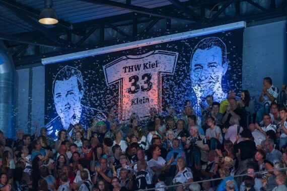 Eine große Menge von Fans versammelt sich in der Halle unter einem Banner mit zwei illustrierten Gesichtern und einem Trikot mit THW Kiel 33 Klein. Konfetti fällt von oben, die Menschen jubeln und machen Fotos.