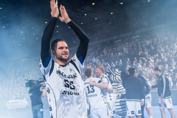 Ein Handballspieler in einem weiß-schwarzen Trikot klatscht mit erhobenen Armen und lächelt inmitten von Rauch auf einem überfüllten Hallenplatz. Teamkollegen und ein Maskottchen stehen in der Nähe, während die Fans im Hintergrund jubeln.