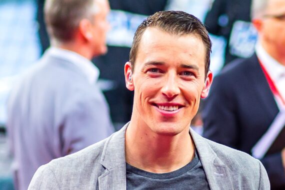 Ein lächelnder Mann mit kurzen braunen Haaren, der einen grauen Blazer über einem dunklen Hemd trägt, steht bei einer Veranstaltung im Mittelpunkt. Mehrere Personen in Geschäftskleidung sind im Hintergrund unscharf.