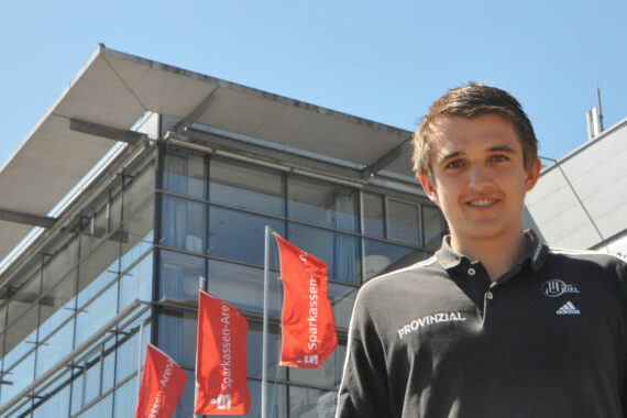 Ein junger Mann in einem schwarzen Poloshirt steht vor einem modernen Glasgebäude mit roten Sparkassen-Arena-Fahnen. Der Himmel ist klar und sonnig.