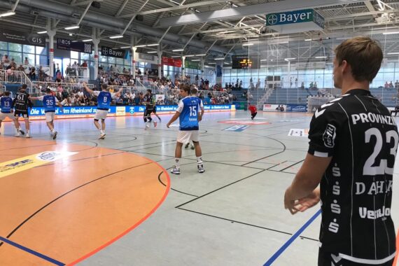 Bei einem Handballspiel in einer Halle sind Spieler in blauen und weißen Trikots auf dem Spielfeld zu sehen. Ein Spieler in einem schwarzen Trikot mit der Nummer 23 bereitet sich darauf vor, von der Seitenlinie aus ins Spiel zu kommen; die Zuschauer füllen die Tribüne.