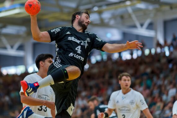 Ein Handballspieler in einem schwarzen Trikot springt in der Luft, um einen Ball zu werfen, während Spieler in weißen Trikots während eines Hallenspiels mit einer unscharfen Menschenmenge im Hintergrund zusehen.