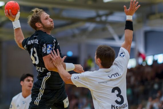 Ein männlicher Handballspieler in einem schwarzen Trikot springt, um den Ball zu werfen, während ein Verteidiger in einem weißen Trikot versucht, ihn zu blockieren. Das Spiel findet in einer Halle mit Zuschauern im Hintergrund statt.