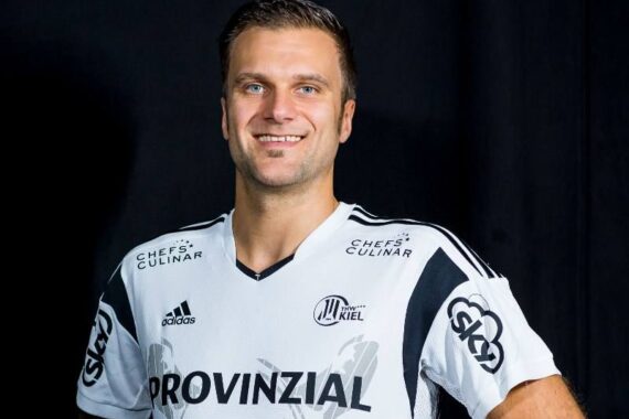 Ein Mann in einem weiß-schwarzen Sporttrikot mit Provinzial- und Sponsorenlogos steht vor einem dunklen Hintergrund und stützt lächelnd die Hände in die Hüften.