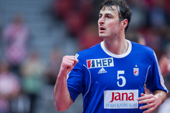 Ein männlicher Handballspieler in einem blauen Trikot mit der Nummer 5 ballt während eines Spiels die Faust, möglicherweise um zu feiern. Der Hintergrund ist unscharf mit roten und grauen Tönen, die an eine Sporthalle erinnern.