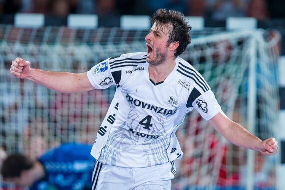 Ein männlicher Handballspieler in einem weißen Trikot mit der Nummer 4 feiert leidenschaftlich mit geballten Fäusten auf einem Hallenplatz, wobei im Hintergrund ein Torwart und ein Tor unscharf zu erkennen sind.