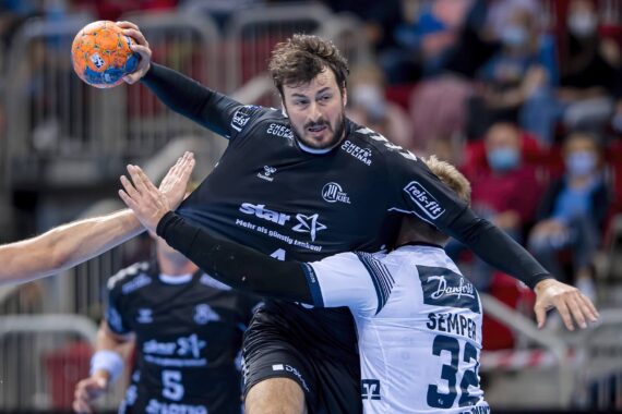 Ein männlicher Handballspieler in einem schwarzen Trikot springt, um den Ball zu werfen, während ein Verteidiger in einem weißen Trikot sein Trikot festhält. Die Aktion findet in einer Halle statt, mit einem unscharfen Publikum im Hintergrund.