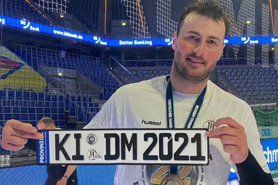 Ein lächelnder Mann mit weißem Hemd und Medaille hält in einer Sporthalle ein Schild, das aussieht wie ein Autokennzeichen mit der Aufschrift KI DM 2021.