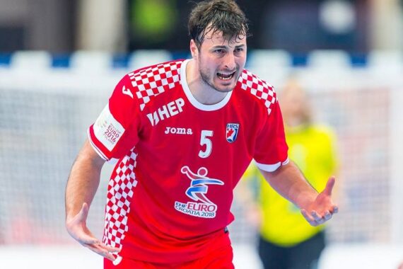 Ein männlicher Handballspieler in einem roten Trikot mit der Nummer 5 gestikuliert leidenschaftlich während eines Spiels. Das Trikot weist ein Muster der kroatischen Flagge auf den Schultern auf. Im Hintergrund ist ein verschwommener Schiedsrichter oder Torwart zu sehen.