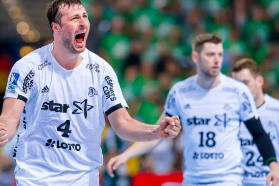 Ein Handballspieler in einem weißen Trikot mit der Nummer 4 feiert leidenschaftlich mit geballten Fäusten, während zwei Mannschaftskameraden hinter ihm stehen. Im Hintergrund sind Zuschauer in grünen Trikots zu sehen.