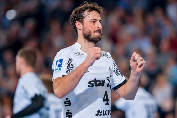 Ein männlicher Handballspieler in einem weißen Trikot mit der Nummer 4 steht auf dem Spielfeld und hebt leicht die Fäuste, während andere Spieler und eine verschwommene Menschenmenge im Hintergrund zu sehen sind.